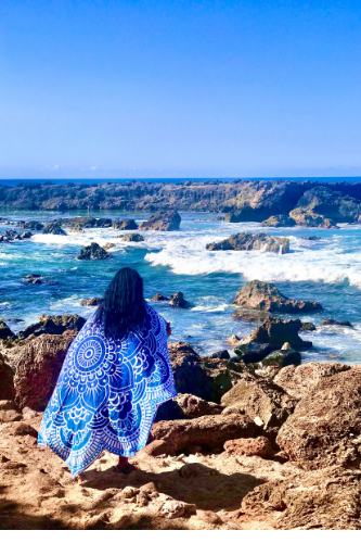 woman standing on stony beach at North Shore Hawaii, looking at Shark's Cove and has a blue mandala beach blanket around her shoulders