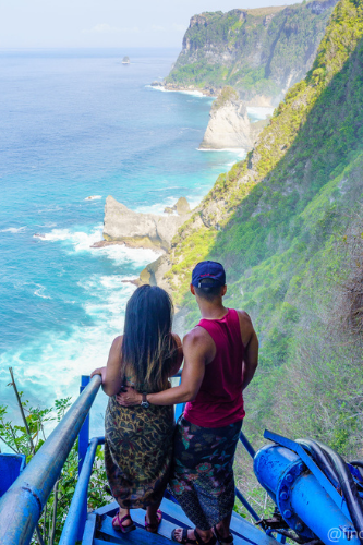 a couple standing on a blue metal steep stairs on the side of a mountain cliff, overlooking the ocean in nusa penida. They are both wearing Teva Sandals.