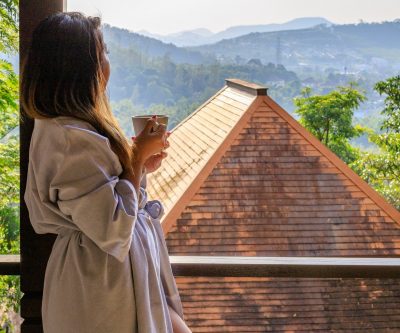 Lifestyle blog. 9 to 5. Woman in bathrobe holding a cup of coffee. She is looking out the window and admiring the trees and mountain views in Phuket Thailand.