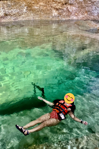 Woman in red bathing suit wearing life vest, floating on a cenote in Tulum, wearing teva sandals and has floating waterproof phone case around her neck