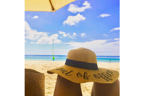 floppy hat with "out of office" print sitting on top of two knees, view of the beach