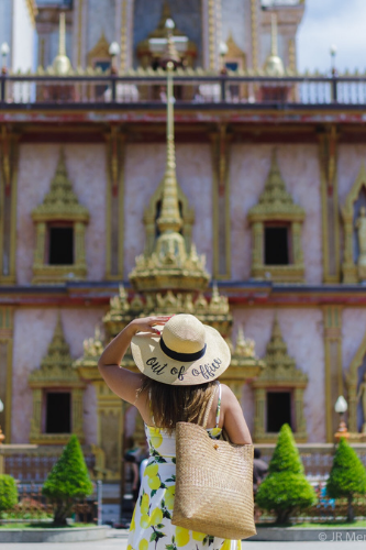 woman wearing floppy hat that has "out of office" print. She is wearing lemon print dress, has a straw bag, and looking at the temple